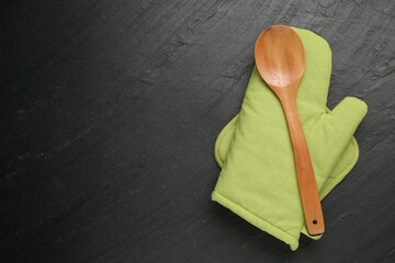 Green oven glove, wooden spoon and potholder on dark textured table, top view. Space for text