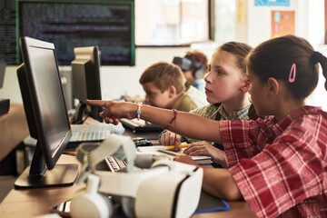 Caucasian girl and Caucasian boy collaborating at computer workstation in classroom, girl pointing at monitor while classmates working in background, technology education concept