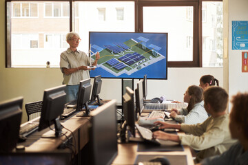 Caucasian senior woman presenting solar panel model to diverse group of young adults in computer...