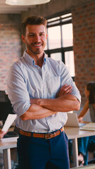 Portrait Of Smiling Businessman Standing In Busy Office With Colleagues Working In Background