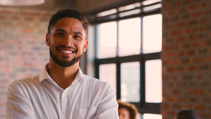Portrait Of Smiling Businessman Standing In Busy Office With Colleagues Working In Background