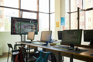 Empty classroom with multiple computer workstations displaying programming code on monitors, modern educational setting with large windows and backpacks on chairs