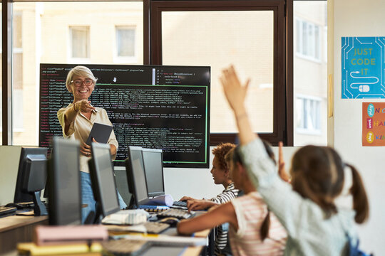Senior Caucasian woman teaching group of diverse children in computer classroom, holding digital tablet while smiling and pointing at students raising hands in front of monitors