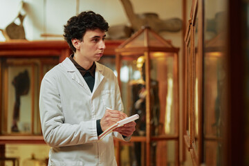 Young adult Caucasian man wearing lab coat standing in museum exhibit area writing notes in notebook while observing display case with scientific specimens