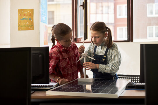 Two Caucasian girls working together assembling wind turbine model on solar panel in classroom, focusing on renewable energy project, both appearing as children around elementary school age