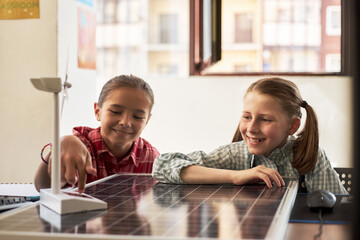 Two Caucasian girls sitting at desk interacting with model wind turbine and solar panel, smiling...