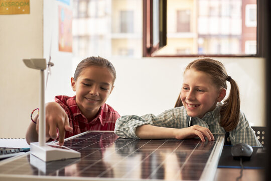 Two Caucasian girls sitting at desk interacting with model wind turbine and solar panel, smiling and engaging in science activity in classroom setting, natural light coming through window