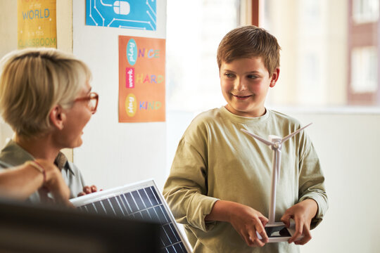 Caucasian boy holding wind turbine model and smiling at middle aged Caucasian woman with short hair, woman holding solar panel, both standing in classroom with educational posters