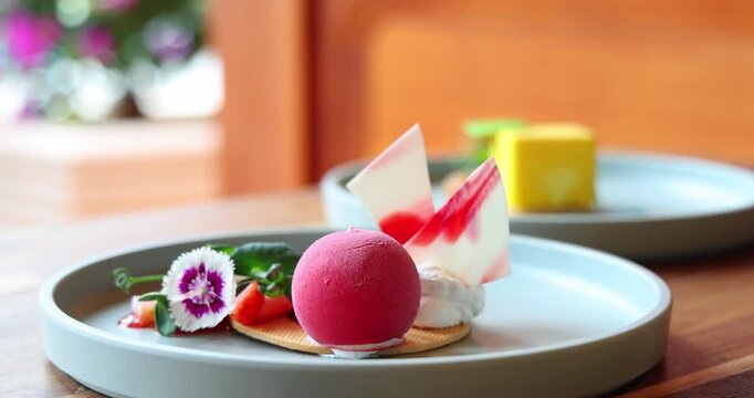 Gourmet dessert plate featuring vibrant raspberry mousse, artistic chocolate shards, fresh fruits, garnished with edible flowers on wooden table. Elegant culinary presentation in natural setting.