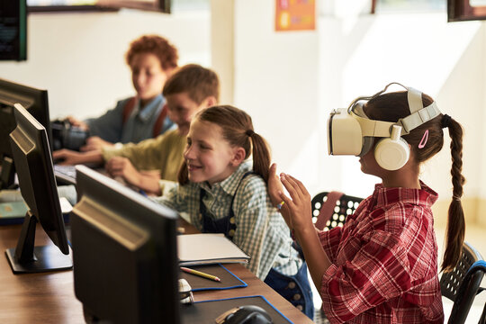 Group of Caucasian and Black children sitting at desks using computers in classroom, girl wearing virtual reality headset interacting with digital content, classmates watching and smiling
