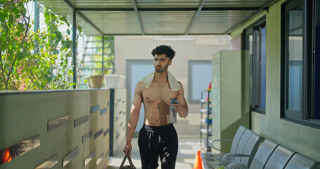 Indian gen z young adult male man standing indoor hall way hold bottle opening drink fresh water Asian well being boy guy take break after sports workout match game spend time inside corridor area
