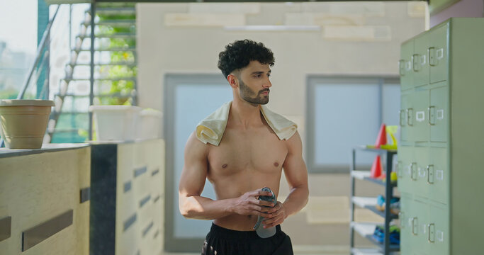 Indian gen z young adult male man standing indoor hall way hold bottle opening drink fresh water Asian well being boy guy take break after sports workout match game spend time inside corridor area - Powered by Adobe