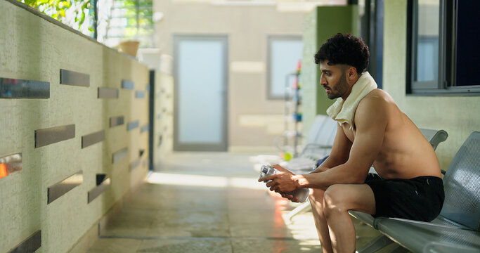 Indian gen z young adult male man guy sitting on chair in hallway hold bottle. Asian relax boy take break after sports workout spend gym time look sad upset outside corridor area