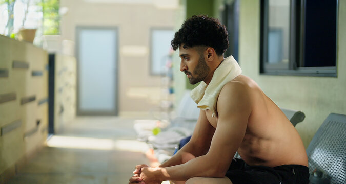 Indian gen z young adult male man guy sitting on chair in hallway. Asian relax boy take break after sports workout spend gym time look sad upset outside corridor area - Powered by Adobe