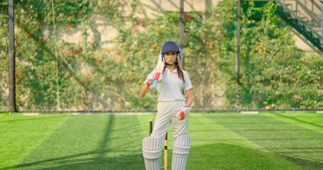 Obraz premium Indian young adult gen z sports woman lady standing at outdoor ground day wear white uniform and helmet look camera Asian one player female girl hold bat staring cam play with ball do casual pose