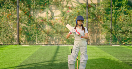 Indian young adult gen z sports woman lady standing at outdoor ground day wear white uniform and helmet look camera. Asian female girl player hold bat staring cam play do casual pose