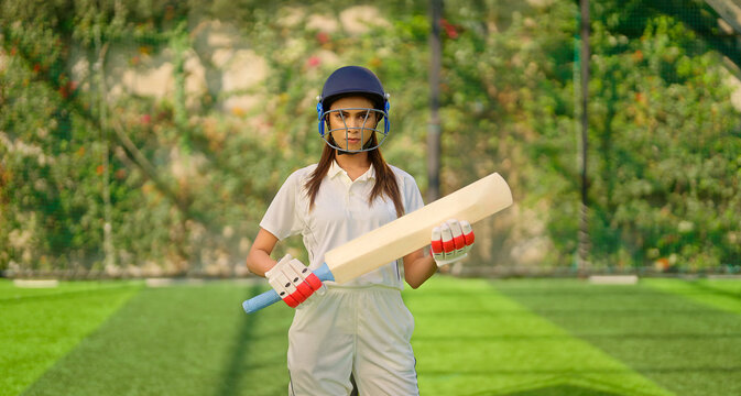 Indian young adult gen z sports woman lady standing at outdoor ground day wear white uniform and helmet look camera Asian one player female girl hold bat staring cam play with ball do casual pose	 - Powered by Adobe