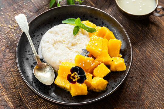 Thai mango sticky rice with coconut milk, garnished with edible flowers on wooden background close up