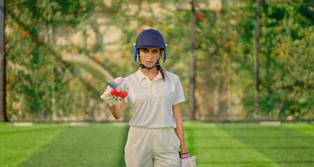 Indian gen z athlete sports woman lady standing at outdoor ground day wear white uniform and helmet look camera Asian one cricket player female girl hold bat staring cam play with ball do casual pose 