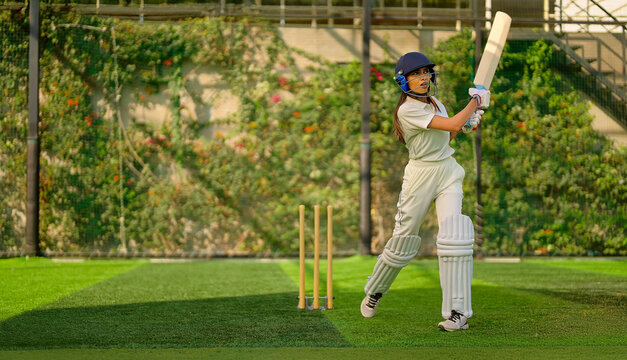 Indian young adult gen z sports woman lady standing hold bat play learn cricket match do practice to hit ball at garden angry teen female girl player wear white uniform leg guard hand gloves day time