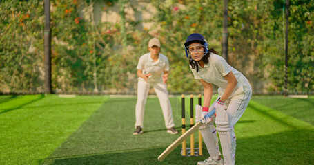 Fototapeta premium Indian gen z young adult sports woman girl standing at outdoor match play ground wear gloves white uniform and protective helmet Asian athlete cricket player female lady hold bat do practice day time