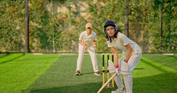 Indian gen z young adult sports woman girl standing at outdoor match play ground wear gloves white uniform and protective helmet Asian athlete cricket player female lady hold bat do practice day time - Powered by Adobe