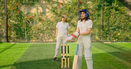 Fototapeta premium Indian gen z young adult sports girl at match play ground wear gloves white uniform leg guard and helmet Asian two cricket player female lady standing hold bat do casual pose ready for match day