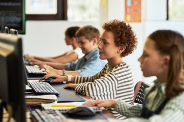 Group of multiethnic children sitting at desks using desktop computers in classroom, focusing on...