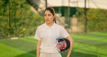 Indian gen z relax sports teen age girl woman standing outdoor play ground wear white uniform hold protective helmet and hand gloves Asian cricket player female take break rest time at match ground