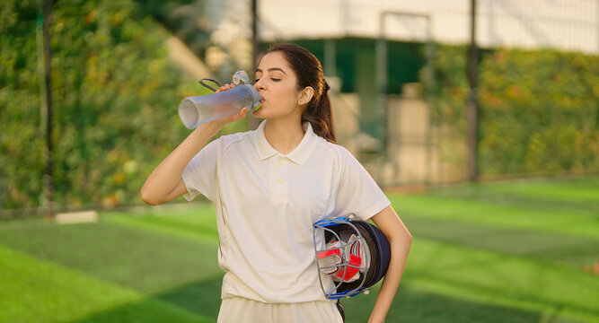 Indian gen z athlete sports teen age girl woman standing outdoor play ground wear white uniform hold protective helmet and hand glove. Asian cricket player female take break time drink water at match	