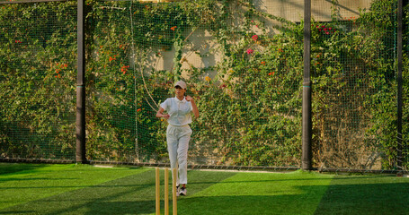Indian young adult gen z bowler woman lady standing at match ground wear white uniform with cap. Asian teen age female girl running play cricket learn game do practice to hit wicket throw ball place