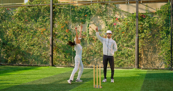 Indian young adult gen z bowler woman standing outdoor match play ground wear uniform cap. Asian happy female lady throwing ball do practice clap hand umpire man show one hands out signal