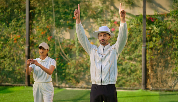 Indian young adult gen z bowler woman standing outdoor match play ground wear uniform cap Asian female lady throwing ball do practice clap hand umpire man show gesture of six with two hands up signal