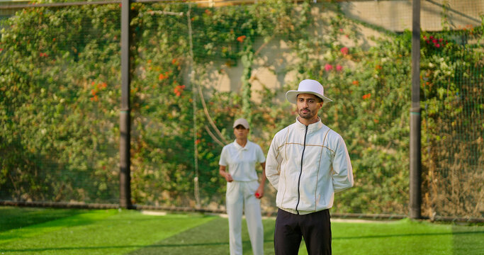 Indian young adult gen z bowler woman standing outdoor match play ground wear uniform cap Asian female lady throwing ball do practice umpire man standing watch player cricket game