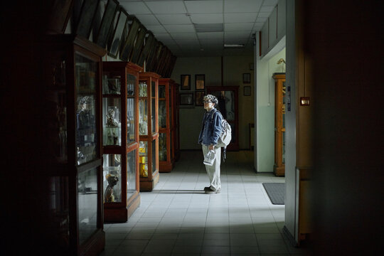 Caucasian middle aged man standing in dimly lit hallway examining glass display cases, holding newspaper and wearing backpack, illuminated by overhead light, appearing thoughtful