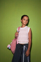 Portrait of Caucasian girl child standing against green wall holding pink backpack with plush toy...