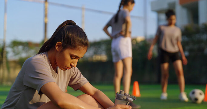 Indian happy athlete woman lady player group play football game kicking soccer ball team outdoor young adult gen z tired girl female sitting take break practice health care hold bottle drink water