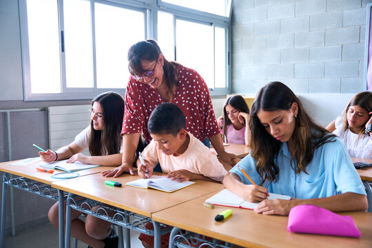 Elementary school teacher assisting students with their schoolwork, guiding a young boy while other children focus on writing in their notebooks at their desks
