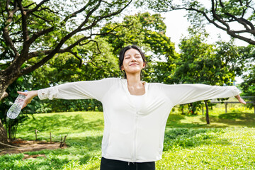 A joyful young asian woman is gracefully stretching her arms outdoors in a lush, expansive green...
