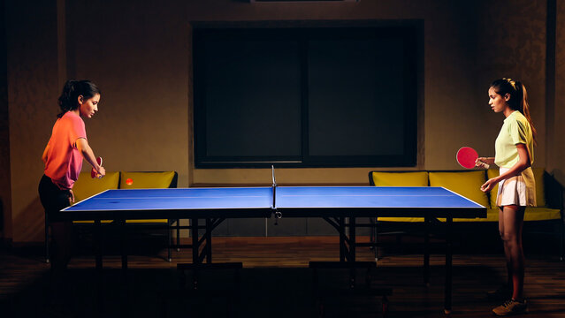 Indian happy young adult gen z two athlete player woman hold racket play table tennis passing ball indoor dark room. Asian sporty girl lady do sports training enjoy ping pong game have fun joy