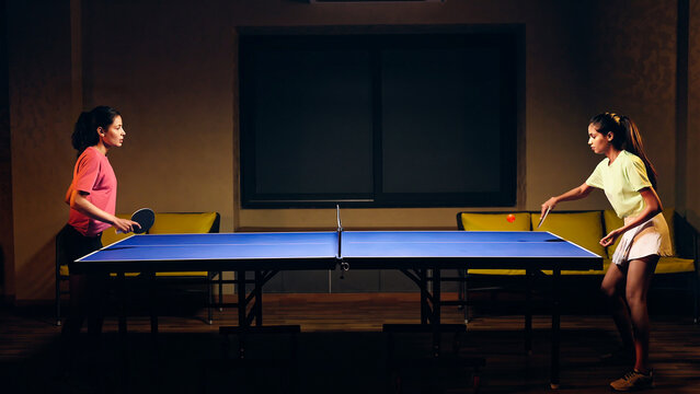 Indian happy young adult gen z two athlete player woman hold racket play table tennis passing ball indoor dark room. Asian sporty girl lady do sports training enjoy ping pong game have fun joy