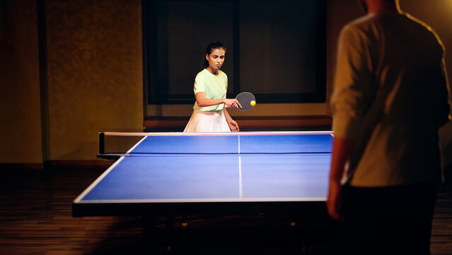 Indian happy young adult gen z athlete player woman hold racket play table tennis male man passing ball indoor dark room Asian sporty girl lady do sports training enjoy ping pong game have fun joy	