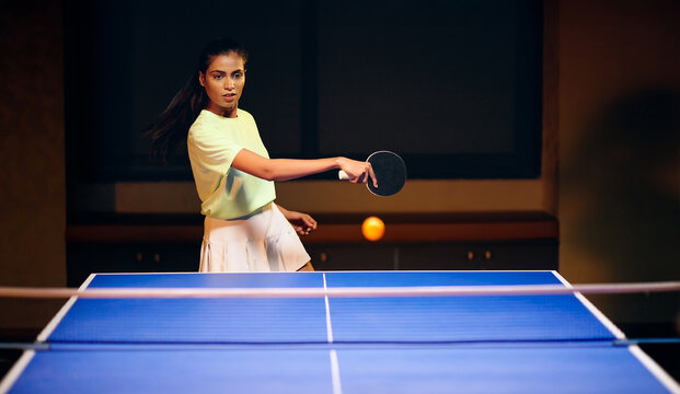 Indian young adult gen z athlete player woman hold racket play table tennis passing ball indoor dark room. Asian sporty girl lady do sports training enjoy ping pong game have fun joy	