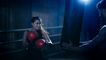Indian young adult gen z angry fit boxer woman lady wear gloves do hand fist boxing punch beat on bag indoor dark room place modern sports female girl learn self defense martial art skill