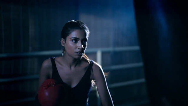 Indian young adult gen z angry fit boxer woman lady wear gloves do hand fist boxing punch beat on bag indoor dark room place modern sports female girl learn self defense martial art skill - Powered by Adobe