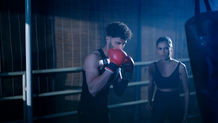 Indian young adult gen z angry fit boxer man guy wear gloves do hand fist boxing punch beat hit on bag indoor dark class room place modern sports smart male boy learn self defense martial art skill