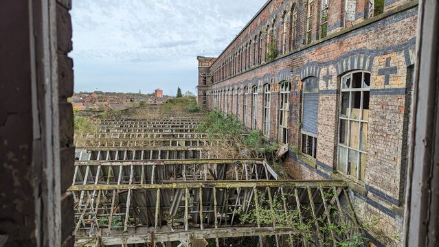 Abandoned mill, derelict historical architecture. Collapsing factory roof, window frame overlooking rooftop ruins. Nature reclaiming crumbling post-apocalyptic industrial building. Brick framing decay