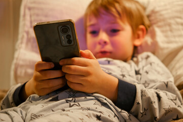 Cute kid boy in pajamas lying in bed watching smartphone, smiling. Children's screen time. Child plays games using phone at the evening or night. Social media addiction. Childhood, modern technology