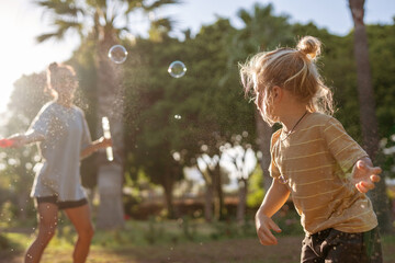 Fototapeta premium Curious blond-haired little boy watching soap bubbles floating in the air on a sunny afternoon in a park, with soft light, green trees, and a woman playing in the background