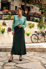 Elegant woman in a green buttoned cardigan and long satin skirt posing outdoors on a sunny day,...